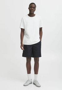 T-shirt en coton blanc, shorts marine, baskets grises et chaussettes blanches de type crew. Le modèle se tient devant un fond uni. Coupe décontractée.