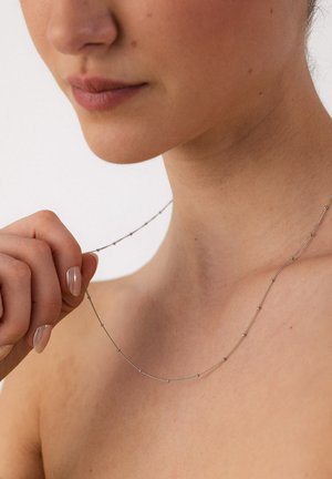 Woman holding a delicate silver beaded necklace near her neck, showing natural skin and neutral-toned lips, against a plain background.