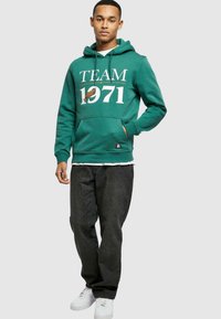 Zöld kapucnis felső "TEAM 1971" felirattal és logónyomattal, kenguruzsebb, bordázott mandzsetták, sötét farmernadrággal és fehér tornacipővel párosítva. Lazán illeszkedő.