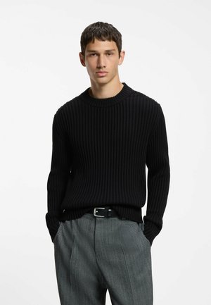 Ung mand med kort mørkt hår iført en sort ribstrikket sweater og grå mønstrede bukser med hænderne i lommerne mod en ensfarvet hvid baggrund.
