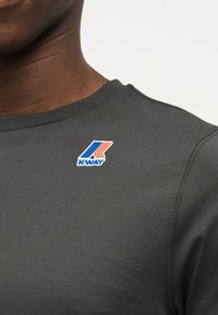 Κοντινό πλάνο ενός σκούρου γκρι πουκαμίσου με ένα μικρό μπλε, πορτοκαλί και λευκό λογότυπο K-Way κοντά στον γιακά στο αριστερό μέρος του στήθους.