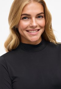 Crna majica s visokim ovratnikom, glatke teksture, prilagođenog dizajna i dugih rukava. Model prikazuje suptilan osmijeh, ističući izrez i tkaninu.