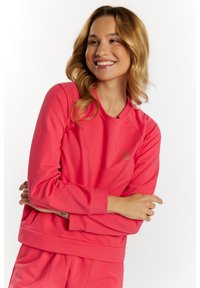 Koraloowy sweter z okrągłym dekoltem, długimi rękawami raglanowymi i ściągaczami. Posiada subtelny detal z logo na piersi.