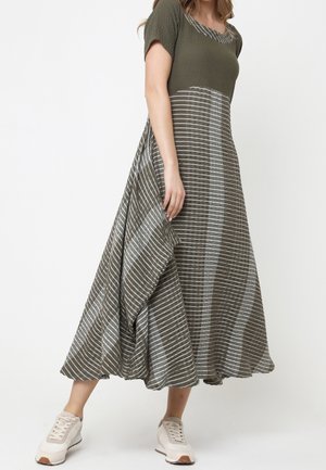 Vrouw draagt een olijfgroen en grijs gestreepte maxi-jurk met korte mouwen, gecombineerd met witte sneakers, staat met één been naar voren.