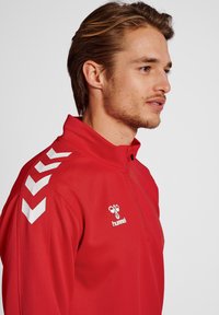 Giacca sportiva rossa con colletto alto, motivi a V bianchi sulle maniche e logo "Hummel". Tessuto liscio, design sportivo.