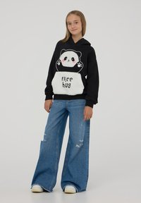 Czarna bluza z kapturem z motywem pandy i napisem "DARMOWY uścisk", w zestawieniu z szerokimi niebieskimi dżinsami. Miękki materiał i puszysty biały akcent z przodu.