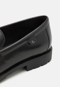 Mustmustelised nahast loafersid sujuva viimistlusega, ümar varvas, tume must kummist tald ja reljeefne logo detail küljel.