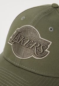 Olivově zelená čepice s vyvýšeným, vyšitým logem "Lakers". Má hladkou texturu látky a strukturovaný tvar s detaily šití.