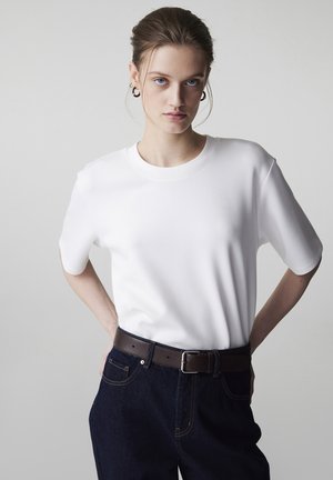 Młoda kobieta o niebieskich oczach i jasnobrązowych włosach związanych do tyłu, ubrana w prostą białą koszulkę z krótkim rękawem, ciemne jeansy i brązowy pasek.