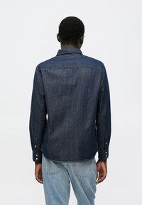 Tamsios džinsinės medžiagos marškiniai su sagomis, ilgomis rankovėmis, klasikiniais apykakle ir dviejų sagų rankogaliais. Lygus audinys su subtiliais spalvos atspalvių pokyčiais visoje marškinių medžiagoje.