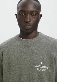 Pelēka adīta džemperis ar apaļu izgriezumu. Kreisajā krūšu daļā ir balta izšūta teksta "LA FLAMME ROUGE". Mīksta tekstūra.