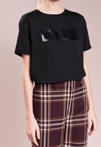 Czarny t-shirt z krótkim rękawem z błyszczącym czarnym logo "HUGO" na prostokątnej naszywce. Do tego spodnie w kratę w kolorze burgundu i kremu.