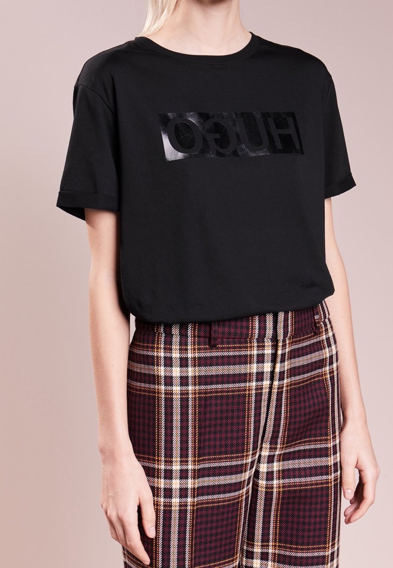 Czarny t-shirt z krótkim rękawem z błyszczącym czarnym logo "HUGO" na prostokątnej naszywce. Do tego spodnie w kratę w kolorze burgundu i kremu.