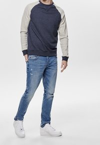 Granatowa bluza z szarymi, raglanowymi rękawami, zestawiona z przetartymi, jasnoniebieskimi dżinsami i białymi tenisówkami. Casualowy styl, detale z teksturowanego materiału.