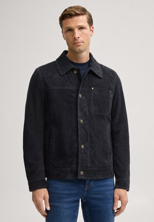 Mann trägt eine schwarze, geknöpfte Jacke mit Brusttasche und blaue Jeans, steht vor einem schlichten hellen Hintergrund.