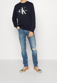 Person klädd i en svart Calvin Klein Jeans sweatshirt, blå slitna skinny jeans och orange sneakers, stående mot en helt vit bakgrund.