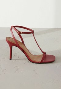 Sandal con tacco alto in pelle rossa, con sottili cinturini, design a forma di T e fibbia. Texture liscia e soletta beige con dettagli minimi.