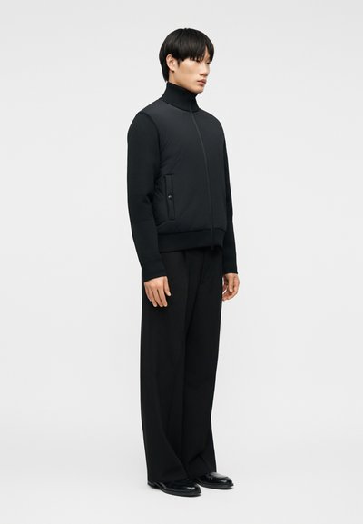 Νεαρός άνδρας όρθιος, φοράει μαύρο μπουφάν με φερμουάρ και τσέπες, φαρδιά μαύρη παντελόνια και μαύρα παπούτσια, κοιτάζοντας ελαφρώς προς τα δεξιά.