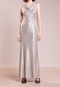 Maxi šaty s kýchacími průramky z metalické stříbrné látky s hladkým povrchem, které mají přiléhavý střih a jsou doplněny černými otevřenými botami.
