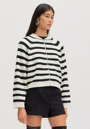 Vrouw die een zwart-wit horizontaal gestreepte sweater met rits draagt, zwarte short en grote zilveren spoel oorbellen tegen een effen achtergrond.