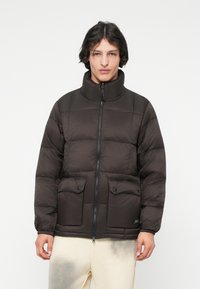 Sötétbarna párnázott dzseki magas gallérral, két elülső hajtókás zsebbel és cipzáras záródással. Simító textúra és téglalap alakú steppelt minta.