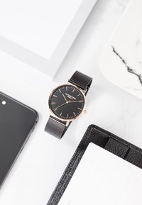 Reloj negro con un diseño minimalista, acentos en oro rosa, correa de malla y marcadores de horas simples en un fondo oscuro. Rodeado de material de oficina y un teléfono.