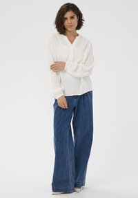 Femme aux cheveux courts et bouclés portant une blouse blanche à boutons et manches bouffantes, un jean large bleu et des chaussures beiges à bouts pointus, se tenant devant un fond blanc.