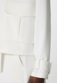 White textured top with a flap pocket, featuring a wide cuff secured with a belt-like strap and matching fabric. Smooth, elevated design.