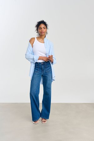 Jonge vrouw met krullend haar, dragend een wit hemdje, een lichtblauw open overhemd, wijde blauwe jeans en open hakken, staand tegen een effen achtergrond.