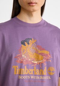 Lilla bomulls T-skjorte med et grafisk trykk av støvler omkranset av røtter, med teksten "Timberland Boots With Roots" under.