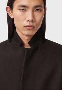 Mustavilla sekoitekangas takki korkealla kauluksella ja sileällä pinnalla, jossa on minimalistischinen muotoilu ja hienovaraisia ompeluyksityiskohtia.