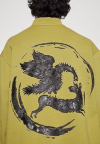 Felpa gialla con un ampio design grafico nero di un uccello e di una creatura, con un accento circolare. Tessuto morbido, vestibilità casual.