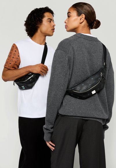 Due modelli che indossano borse a tracolla nere; uno in una maglietta bianca con maniche marroni, l'altro in un maglione grigio e pantaloni scuri, che si fronteggiano.