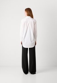 Camicia bianca a maniche lunghe con orlo curvo, polsino con bottone e dettaglio arricciato sul retro, abbinata a pantaloni neri a gamba larga.