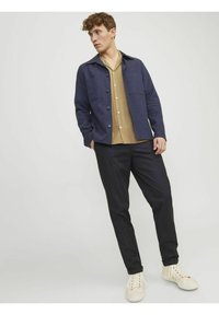 Giacca a camicia blu navy con bottoni, camicia beige con colletto sotto, pantaloni neri affusolati e sneakers bianche, in piedi su uno sfondo neutro.