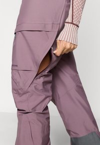 Fialové turistické nohavice s viacerými vreckami na zips, pružným materiálom a kontrastnými sivými doplnkami na spodnej časti nôh. Majú ventilačné otvory.