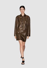 Abito in pelle marrone con colletto, maniche lunghe e bottoni frontali; presenta tasche laterali e una texture liscia. Calzature nere.