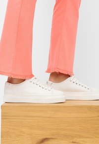 Witte leren sneakers met ronde neuzen, vlakke veters en rubberen zolen. Gepaard met cropped, gescheurde koraalkleurige broekjes die op een houten oppervlak liggen.