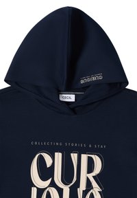 Sweat à capuche bleu marine avec l'étiquette "CECIL", grand texte stylisé "CUR" sur le devant, et petite inscription "CURIOUS NOMAD OF THE SOUL" sur la capuche.