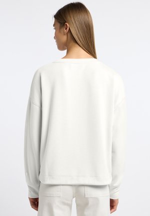 Vrouw met steil lichtbruin haar draagt een losse witte sweatshirt met lange mouwen en witte broek, gezien van achteren tegen een effen achtergrond.