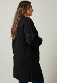 Schwarze Strickjacke mit einem strukturierten Rautenmuster, langen Ärmeln und seitlichen Schlitzen für mehr Bewegungsfreiheit, seitlich präsentiert.