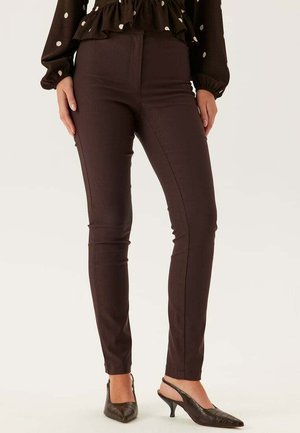 Donna che indossa pantaloni aderenti marrone scuro e décolleté slingback neri con tacco basso, in piedi con le mani rilassate ai lati.