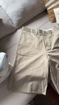 Pantaloni corti color khaki realizzati in un tessuto liscio, con una parte anteriore piatta, passanti per cintura e tasche laterali. La texture è leggermente stropicciata.