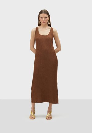 Robe maxi plissée marron avec un décolleté arrondi, présentant une texture lisse. Associée à des sandales à talons dorés et des boucles d'oreilles triangulaires.
