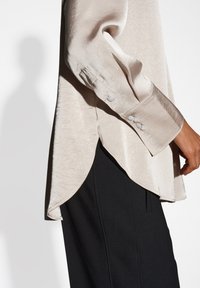 Blusa beige chiaro, testurizzata, con orlo a spacco laterale e polsini abbottonati, abbinata a pantaloni neri. Tessuto liscio con una leggera lucentezza.