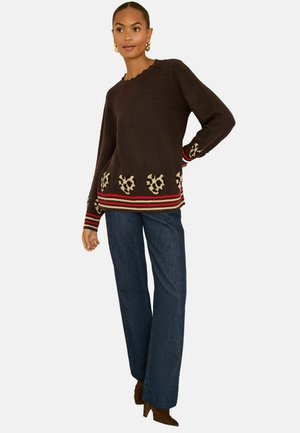 Barna kötött pulóver leopárdmintás díszítéssel és piros csíkkal az alján, szorosan illeszkedő kék farmernadrággal és barna magassarkú csizmával párosítva.