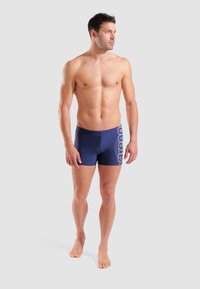 Marineblaue Badehose mit weißem Logo an der Seite, enganliegendes Design, glatte Textur und Oberschenkellänge, aus dehnbarem Material gefertigt.