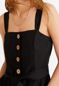Vrouw in een mouwloze zwarte jurk met een verticale rij van vier houten knopen en een dunne gouden ketting. 