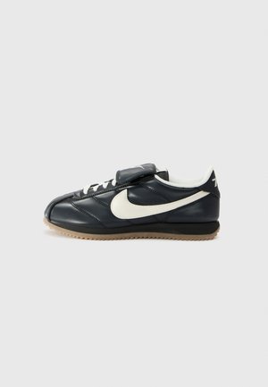 Melna ādas sporta apavi ar baltu swoosh logotipu, auklīšu aizdari, velcro siksniņu un gumijas zoli ar teksturētu rakstu. Parādīts sānu skats.