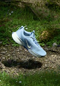 Šviesiai mėlynas sportinis batas su baltu padu ir Nike swoosh, pakabintas virš akmenuoto grunto, aplink jį auga žalia žolė ir mažos gėlės.